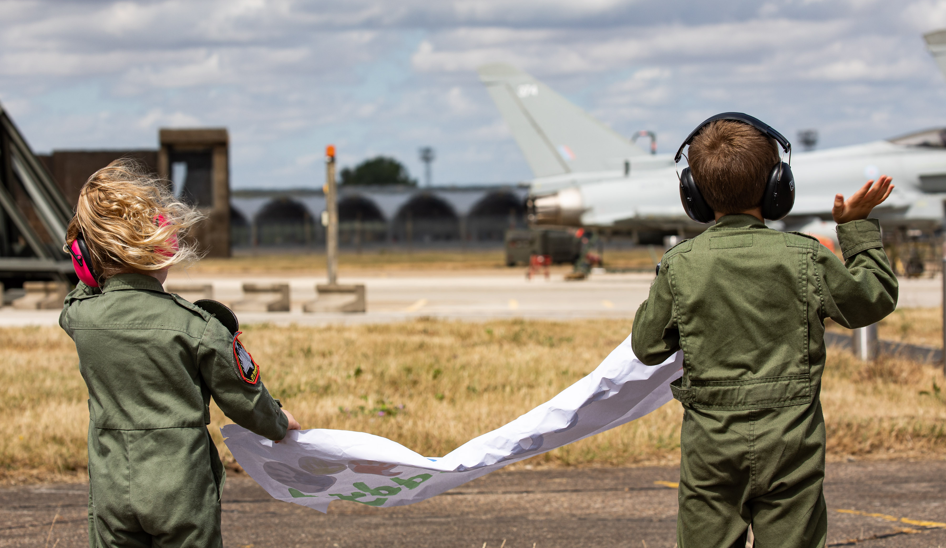 Image shows two children waving on the airfield and holding poster.